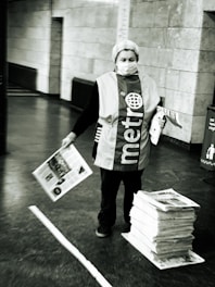 An individual is standing in a public space, wearing a vest with the word 'metro' on it. They are holding newspapers, while a stack of newspapers lies on the ground beside them. The setting appears to be an indoor area with tiled walls and a concrete floor.