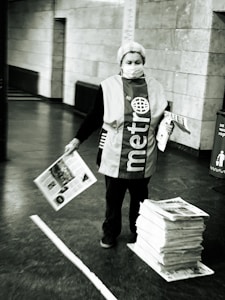An individual is standing in a public space, wearing a vest with the word 'metro' on it. They are holding newspapers, while a stack of newspapers lies on the ground beside them. The setting appears to be an indoor area with tiled walls and a concrete floor.
