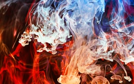 Vibrant swirls of colored smoke intertwine, creating a dynamic and fluid pattern. Blue, red, and orange hues dominate the scene, blending seamlessly to form abstract shapes that evoke a sense of motion and energy.