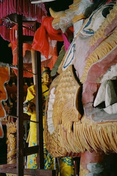 A richly detailed wooden dragon sculpture painted with beige, blue, and red hues dominates the foreground. The intricate carvings and textures highlight the dragon's features. In the background, a figure dressed in a yellow garment with blue patterns is partially visible, accompanied by various draped pink and orange fabrics.