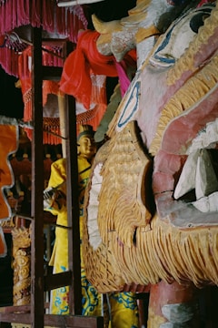 A richly detailed wooden dragon sculpture painted with beige, blue, and red hues dominates the foreground. The intricate carvings and textures highlight the dragon's features. In the background, a figure dressed in a yellow garment with blue patterns is partially visible, accompanied by various draped pink and orange fabrics.