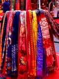 A vibrant display of colorful, intricately patterned and textured hanging scarves, showcasing an array of cultural designs and motifs. The rack is filled with hues ranging from deep blues and vivid reds to bright yellows and soft pastels, creating a visually captivating scene.
