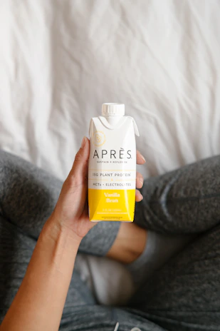 A hand is holding a tetra pak carton of Après Vanilla Bean flavored plant protein drink. The person holding the drink is wearing grey sweatpants and is sitting on a soft, white surface.