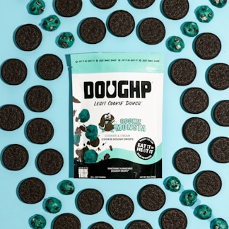 A package of cookie dough is surrounded by chocolate sandwich cookies and blue-colored cookie dough drops on a light blue background. The package features branding and a character designed to look like a cartoon cookie monster. The text indicates the cookie dough is cookies and cream flavored and includes instructions that it can be eaten raw or baked.