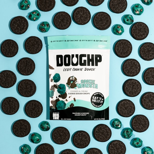 A package of cookie dough is surrounded by chocolate sandwich cookies and blue-colored cookie dough drops on a light blue background. The package features branding and a character designed to look like a cartoon cookie monster. The text indicates the cookie dough is cookies and cream flavored and includes instructions that it can be eaten raw or baked.