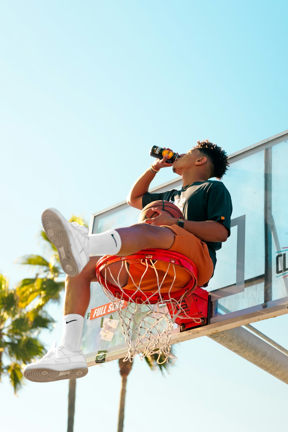 a man sitting on top of a basketball hoop drinking a beer
