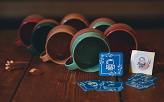 A collection of ceramic mugs lying on their sides with a rustic finish, predominantly in shades of brown, green, and red. In front of the mugs, a series of blue business cards with 'Dilan Pottery' and a small 'Thank you' note are displayed. The scene is set on a wooden surface, giving a cozy, handcrafted vibe. Small decorative wooden leaves and a flower are placed near the mugs.