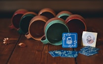 A collection of ceramic mugs lying on their sides with a rustic finish, predominantly in shades of brown, green, and red. In front of the mugs, a series of blue business cards with 'Dilan Pottery' and a small 'Thank you' note are displayed. The scene is set on a wooden surface, giving a cozy, handcrafted vibe. Small decorative wooden leaves and a flower are placed near the mugs.