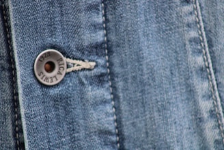 Close-up view of a denim fabric featuring detailed stitching along a seam and a metallic button embossed with the words '1928 RICA LEVI'S'. The weave of the denim fabric is clearly visible, highlighting the texture and the faded blue coloration.