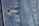 Close-up view of a denim fabric featuring detailed stitching along a seam and a metallic button embossed with the words '1928 RICA LEVI'S'. The weave of the denim fabric is clearly visible, highlighting the texture and the faded blue coloration.