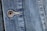 Close-up view of a denim fabric featuring detailed stitching along a seam and a metallic button embossed with the words '1928 RICA LEVI'S'. The weave of the denim fabric is clearly visible, highlighting the texture and the faded blue coloration.