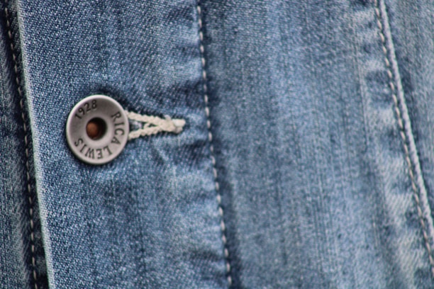 Close-up view of a denim fabric featuring detailed stitching along a seam and a metallic button embossed with the words '1928 RICA LEVI'S'. The weave of the denim fabric is clearly visible, highlighting the texture and the faded blue coloration.