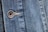 Close-up view of a denim fabric featuring detailed stitching along a seam and a metallic button embossed with the words '1928 RICA LEVI'S'. The weave of the denim fabric is clearly visible, highlighting the texture and the faded blue coloration.