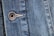 Close-up view of a denim fabric featuring detailed stitching along a seam and a metallic button embossed with the words '1928 RICA LEVI'S'. The weave of the denim fabric is clearly visible, highlighting the texture and the faded blue coloration.