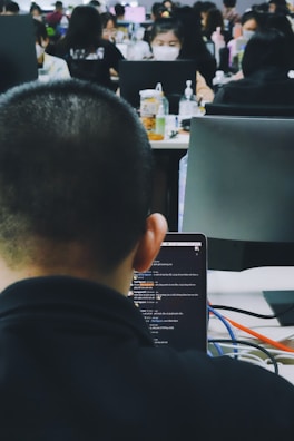 A busy office environment with multiple people working at desks. The focus is on a person using a laptop, with the screen displaying a chat or code editor. Other coworkers are visible in the background, some wearing masks and engaged with their computers.