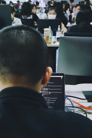 A busy office environment with multiple people working at desks. The focus is on a person using a laptop, with the screen displaying a chat or code editor. Other coworkers are visible in the background, some wearing masks and engaged with their computers.