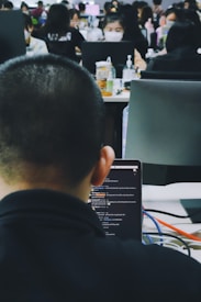 A busy office environment with multiple people working at desks. The focus is on a person using a laptop, with the screen displaying a chat or code editor. Other coworkers are visible in the background, some wearing masks and engaged with their computers.