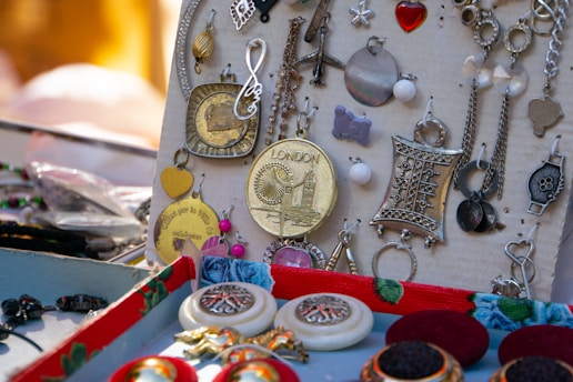 A collection of vintage-style jewelry pieces is displayed on a surface, including various earrings and pendants with intricate designs. Some notable items include a large round pendant with an engraving of Big Ben and the word 'LONDON,' a heart-shaped gold pendant, and ornate silver pieces with floral or geometric patterns.