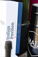 A close-up view of a box with the label 'Italian Pavilion' and some text related to the Mostra Internazionale d'Arte Cinematografica di Venezia, featuring a color-coded design. Beside the box, a dark cylindrical container is partially visible with the text 'MAÌA GARDA DOC' on it.