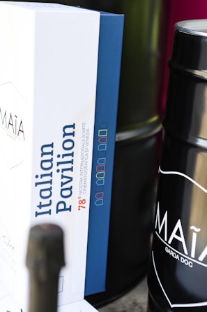 A close-up view of a box with the label 'Italian Pavilion' and some text related to the Mostra Internazionale d'Arte Cinematografica di Venezia, featuring a color-coded design. Beside the box, a dark cylindrical container is partially visible with the text 'MAÌA GARDA DOC' on it.