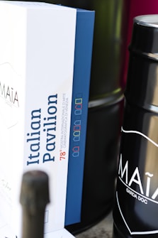 A close-up view of a box with the label 'Italian Pavilion' and some text related to the Mostra Internazionale d'Arte Cinematografica di Venezia, featuring a color-coded design. Beside the box, a dark cylindrical container is partially visible with the text 'MA&Igrave;A GARDA DOC' on it.