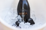 A bottle of sparkling wine labeled 'Maia' nestled in a bucket filled with ice cubes. The bottle appears as a centerpiece, with droplets of condensation indicating its cold temperature.