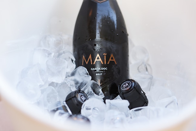 A bottle of sparkling wine labeled 'Maia' nestled in a bucket filled with ice cubes. The bottle appears as a centerpiece, with droplets of condensation indicating its cold temperature.