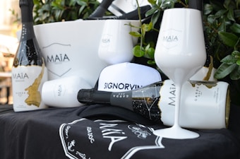 A still life arrangement featuring bottles of Maïa sparkling wine, two white wine glasses with the Maïa logo, a white bucket, and a cap with the word 'Signorvino' on it. The items are set against a backdrop of greenery, creating an elegant and sophisticated atmosphere.