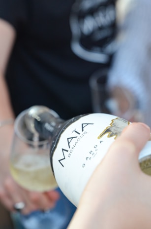 A close-up of a hand pouring wine from a bottle labeled 'Mata Benacus' into a glass. There are blurred figures in the background, suggesting a social setting.