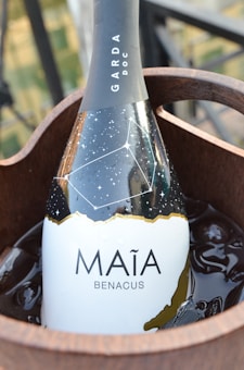 A bottle of wine labeled 'MAÍA BENACUS' rests in a wooden ice bucket. The bottle's design features a constellation pattern over a night sky background. The text 'GARDA DOC' is displayed on the neck of the bottle.