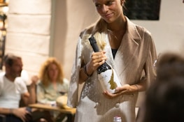 A woman in a shiny, light-colored jacket is holding a bottle of Maia wine. She is standing and smiling, suggesting a social setting. In the background, there are two people sitting at a table, both blurred out. The setting appears to be an indoor dining environment with soft lighting.