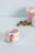 A white ceramic cup features a whimsical snowman design with a red hat and scarf, yellow button decoration, surrounded by a festive star-patterned band. In the blurred background, a pink gift box with a ribbon and green leaves with red berries add to the holiday theme.