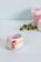 A white ceramic cup features a whimsical snowman design with a red hat and scarf, yellow button decoration, surrounded by a festive star-patterned band. In the blurred background, a pink gift box with a ribbon and green leaves with red berries add to the holiday theme.
