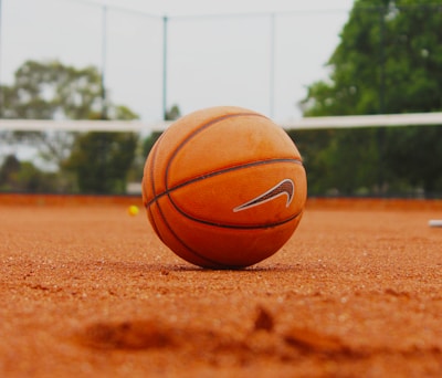 An orange basketball is placed on a clay tennis court. In the background, there is a blurred view of tennis court elements, including the net and surrounding greenery.