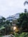Modern apartment buildings with curved glass facades are partially obscured by dense greenery and trees, creating a harmonious blend of architecture and nature. In the foreground, a warmly lit street lamp stands out against the surrounding foliage, adding a cozy element to the scene.