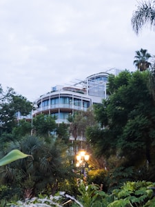 Modern apartment buildings with curved glass facades are partially obscured by dense greenery and trees, creating a harmonious blend of architecture and nature. In the foreground, a warmly lit street lamp stands out against the surrounding foliage, adding a cozy element to the scene.