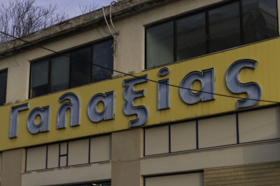 A building facade with a large yellow sign displaying Greek letters in blue. The sign is set against a pale yellow panel. The building features large windows with some showing reflections, and the upper portion has exposed concrete with some vegetation growing over the edge. Overhead wires are visible, and the overall scene suggests an urban setting.