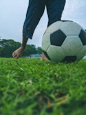 A person stands barefoot on a grassy field, gently nudging a soccer ball with one foot. The lower legs and feet are visible, and the background features green trees and a cloudy sky.