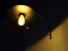 A single glowing light bulb is encased in a circular metal fixture, casting a warm yellow light. The fixture is mounted on a dark wall, with visible wiring running to the right side.
