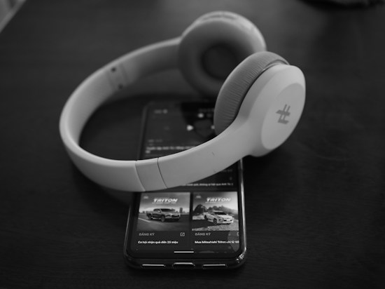 A pair of over-ear headphones rests on top of a smartphone displaying an advertisement for a vehicle, with both items set against a dark, smooth surface. The scene creates a minimalist and modern aesthetic.