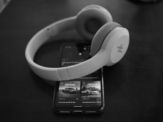 A pair of over-ear headphones rests on top of a smartphone displaying an advertisement for a vehicle, with both items set against a dark, smooth surface. The scene creates a minimalist and modern aesthetic.