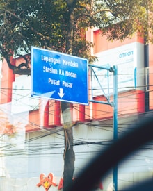 A blue road sign with white text is mounted on a metal pole next to a tree. The sign points downwards, listing destinations including Lapangan Merdeka, Stasiun KA Medan, and Pusat Pasar. The background includes a partial view of a modern building with red and white structures, as well as visible power lines and advertisements.