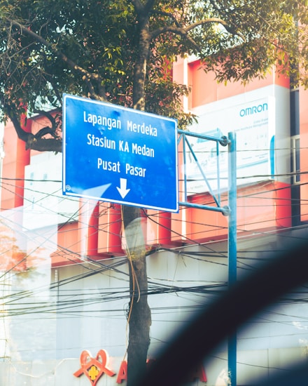 A blue road sign with white text is mounted on a metal pole next to a tree. The sign points downwards, listing destinations including Lapangan Merdeka, Stasiun KA Medan, and Pusat Pasar. The background includes a partial view of a modern building with red and white structures, as well as visible power lines and advertisements.