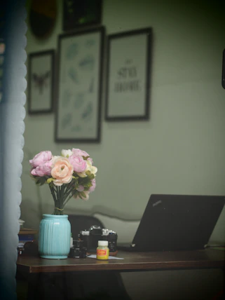 A cozy workspace with a laptop, a cup of coffee, and a vase of fresh flowers beside a window.