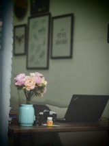 A workspace featuring a laptop and a vase of pastel-colored flowers on a wooden desk. The backdrop includes several minimalistic art frames on the wall. Various small items, such as a pill bottle and some camera lenses, are scattered across the desk.