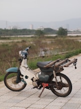 An old, weathered motorcycle with a rustic appearance is parked on a stone pathway. The bike appears to be missing parts and has a makeshift platform attached to the back. In the background, there is a blurred view of a cityscape with buildings and a mountain range under a cloudy sky. Surrounding the pathway are patches of grass and shrubs.