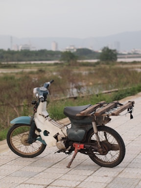 An old, weathered motorcycle with a rustic appearance is parked on a stone pathway. The bike appears to be missing parts and has a makeshift platform attached to the back. In the background, there is a blurred view of a cityscape with buildings and a mountain range under a cloudy sky. Surrounding the pathway are patches of grass and shrubs.