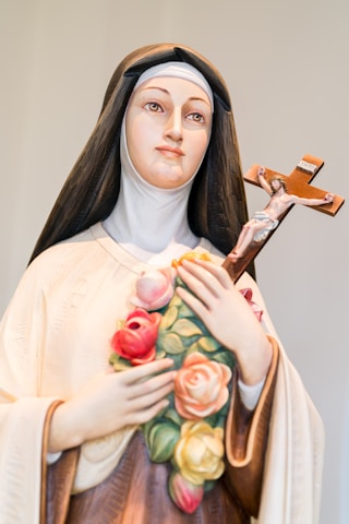 A statue of a woman wearing a veil and a habit, holding a crucifix in one hand and a bouquet of colorful roses in the other. The expression is serene and contemplative.