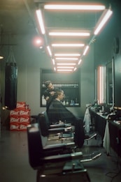 A barbershop with a modern aesthetic featuring a row of barber chairs under linear overhead lighting. A barber is seen styling a customer's hair. Shelves in the background are stocked with grooming products, and a stack of Budweiser boxes is visible on the floor.