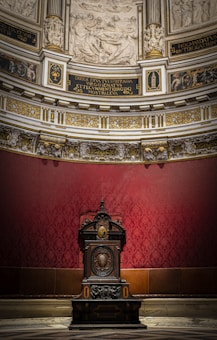 An ornate wooden pulpit or chair is centrally positioned against a richly decorated red wall. The upper section of the image features detailed architectural elements, including intricate carvings and religious artwork. The ceiling area is adorned with Latin inscriptions and gold embellishments, giving the space a regal and sacred atmosphere.
